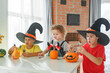 © Olga Gimaeva - Three real siblings two brothers and sister making together jack o lantern at their home white kitchen, cutting real pumpkins and playing with paper black ghosts, lovely vibe of halloween
