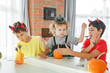 © Olga Gimaeva - Three real siblings two brothers and sister making together jack o lantern at their home white kitchen, cutting real pumpkins and playing with paper black ghosts, lovely vibe of halloween