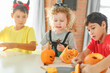 © Olga Gimaeva - Three real siblings two brothers and sister making together jack o lantern at their home white kitchen, cutting real pumpkins and playing with paper black ghosts, lovely vibe of halloween
