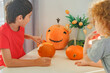 © Olga Gimaeva - Three real happy siblings two brothers and sister cutting together from real natural pumpkin jack o lantern, while playing with paper decorations of ghosts, children wearing with witch hats, halloween