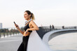 © BGStock72 - Young woman enjoying a call while taking a break from her jog at a tranquil riverside promenade during a sunny afternoon