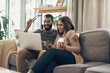 © N F/peopleimages.com - Laptop, coffee and couple on sofa in apartment planning cost, finance and budget for vacation. Happy, computer and people in living room booking accommodation online for travel, holiday or trip.