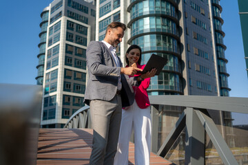 Wall Mural - Smiling Latin Hispanic mature adult businesspeople using pc digital computer. Laughing caucasian colleagues, checking the company budget on their laptop in front of glass building exterior.	