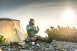© XArt - Stylish bearded man wearing down jacket sitting beside his tent in front of a mountain peak grinding coffee