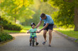 © fotoduets - Young adult father teaching happy beautiful little boy to ride on first bike without pedals on sidewalk at city park. Learning to keep balance. Warm summer day. Cute 2 years old toddler. Front view.