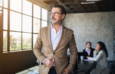 Fototapete - A well-dressed businessman stands by the window in a contemporary office, engaging in a discussion. Two colleagues seated at a table appear focused and attentive during the meeting.