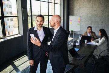 Fototapete - Two men in suits are engaged in a discussion while looking at a smartphone. In the background, colleagues are working at a large table in a stylish office space.
