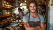 © M_H_L - A smiling male barista stands behind the counter of his cafe with his arms crossed.