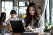 © nikolettamuhari - An Asian woman is working on a laptop while sitting at a table with a digital...