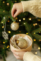 Wall Mural - Decorating christmas tree with vintage golden baubles. Hands in cozy sweater holding stylish Christmas ornaments on background of christmas tree with golden lights. Merry Christmas