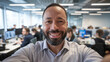 © Tom - Smiling middle-aged Black man takes a selfie in a modern office filled with colleagues working at their desks.
