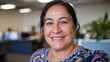 © Tom - Smiling middle-aged Hispanic woman in an office setting, with a colorful floral blouse, exuding warmth and positivity.