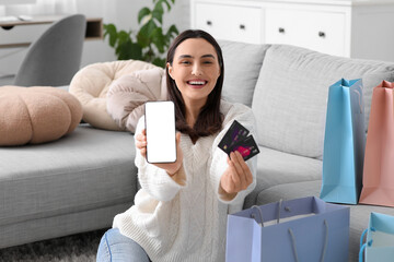 Wall Mural - Happy young woman with mobile phone, credit card and shopping bags sitting on floor at home. Black Friday sale