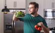 © Volodymyr - Taking care of health. Handsome man in casual t-shirt standing in the kitchen at home. Young man cooking vegetables in the kitchen. Dieting man with bowl of salad. Green salad healthy food concept.