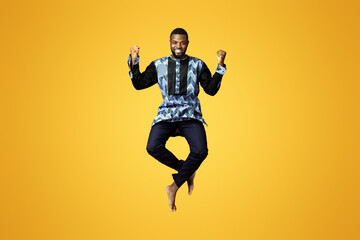 Wall Mural - Emotional black guy showing happiness over pink studio background. Smiling african american man in colorful traditional clothes jumping up and posing, raising fists up, expressing emotions