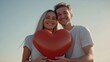 © Daria Lukoiko - A Caucasian couple holding a large heart together, smiling warmly at each other. Soft natural light and a simple sky background, an intimate, joyful atmosphere. Happy Valentine’s day