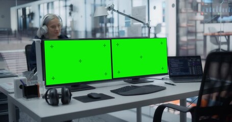 Software Developer Office Workplace with a Desk with Multiple Computer Displays Showing Green Screen Chroma Key Mock Up Templates on Monitors. Young Female Working Behind a Desk in the Background