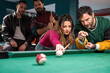 © Lomb - Female pool player aiming white ball while friends watching competitive billiard match during social gathering