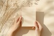 © AiHRG Design - A hand gently cradles a blank postcard mockup in soft beige tones, with the warm, muted background adding a touch of tranquility. The light creates delicate shadows around the scene.