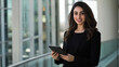 © Maksym - A young Middle Eastern businesswoman dressed in formal attire, standing in a modern office plaza, checking her tablet and smiling, with urban architecture behind her.
