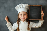 Cooking, culinary and profession concept - happy smiling little girl in chef's toque and jacket with black chalkboard over grey background