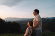 © troyanphoto - A father embraces his daughter as they enjoy the scenic view of hills and mountains under an evening sky.