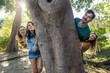 © Lomb - Young diverse friends playing hide and seek behind large tree trunk enjoying playful outdoor activity in sunny park