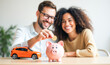 © Radorog. - a happy young couple throws a coin into a piggy bank, next to it is a model of a car, a concept of savings for the purchase of real estate