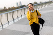 © BGStock72 - A joyful woman in a yellow sweater enjoys a phone call while walking along a riverside promenade on a sunny afternoon