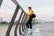© BGStock72 - Young woman in a yellow jacket checks her phone while strolling along a scenic riverside promenade during a calm afternoon