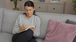 © Krakenimages.com - A young hispanic woman sits comfortably on a living room couch while writing in a notebook, in an inviting indoor home setting featuring a cozy atmosphere and attractive decor.