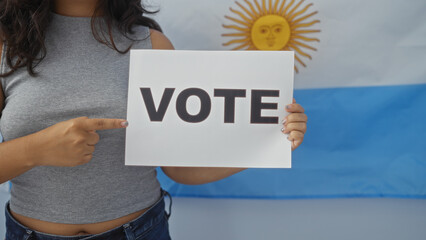 Wall Mural - Young hispanic woman in an electoral college room pointing to a vote sign with an argentinian flag in the background, emphasizing the importance of voting.