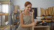 © Krakenimages.com - Redhead woman in a bakery checking her tablet while talking on the phone surrounded by various baked goods and pastries in a cozy indoor shop