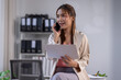 © David - Cheerful young Asian woman discusses work on a phone call in a modern office, holding documents.