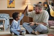 © Seventyfour - Little smiling Black boy giving high five to joyful adoptive father, while celebrating winning at mobile phone game sitting on floor in kids room