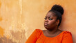 © kudesniza - Pensive black woman in orange shirt leaning against a textured wall
