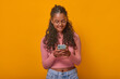 © antianti - Cheerful young Indian woman with curly hair immersed in her smartphone, showcasing a relaxed vibe against a bold yellow wall. Her joyful expression illuminates the lively atmosphere around her.