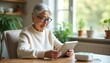 © irissca - A senior woman enjoys reading on a tablet while sitting at a bright table surrounded by plants and natural light during the morning