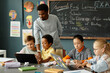 © Seventyfour - Diverse group of primary school children using laptops in pairs to study sitting at large desk in classroom while African American male teacher helping kids working on computers, copy space