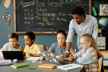 Modern male teacher of African American ethnicity helping young girls with working on laptop in class while children study in pairs at large table in classroom, copy space