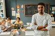 © Seventyfour - Portrait of confident African American male teacher smiling at camera posing with arms crossed in primary school classroom, copy space