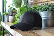 © Gentl - Black baseball cap placed on a wooden surface with potted green plants and flowers in the background. Indoor setting with natural light highlighting the texture and details of the cap.