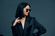 © SHOTPRIME STUDIO - Asian woman in black suit and sunglasses striking a confident pose against a gray background, showcasing elegance and style