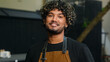 © Yuliia - Happy smiling joyful young Indian Arabian man cafe worker in apron male guy waiter cleaner look distance thinking turning looking at camera friendly wide smile portrait outside city street restaurant