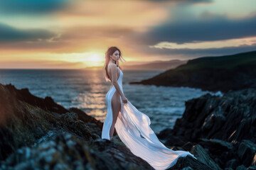 Wall Mural - Woman in flowing white dress gazing at sunset on rocky coastal landscape.