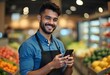 © Priyanka - A young Hispanic man in his 20s with short dark hair wearing a blue shirt and apron, smiling and holding a smartphone in a grocery store or market setting with shelves of produce in the background