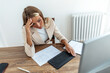 © Jelena Stanojkovic - Exhausted woman having a headache in home office. Creative woman working at office desk with spectacles on head feeling tired. Stressed young woman feeling pain while overworking on desktop computer.