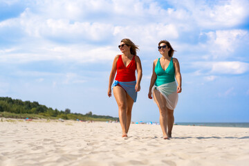 Wall Mural - Two beautiful young females wearing one-piece bathing suits and pareo walking on white sandy beach in summertime. Front view.