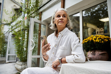 A mature woman sits thoughtfully, holding a smartphone while surrounded by vibrant flowers.