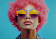 © Amazing Kingdom - A stylish young woman with vibrant pink curls wears colorful round sunglasses against a bright blue background in an indoor setting